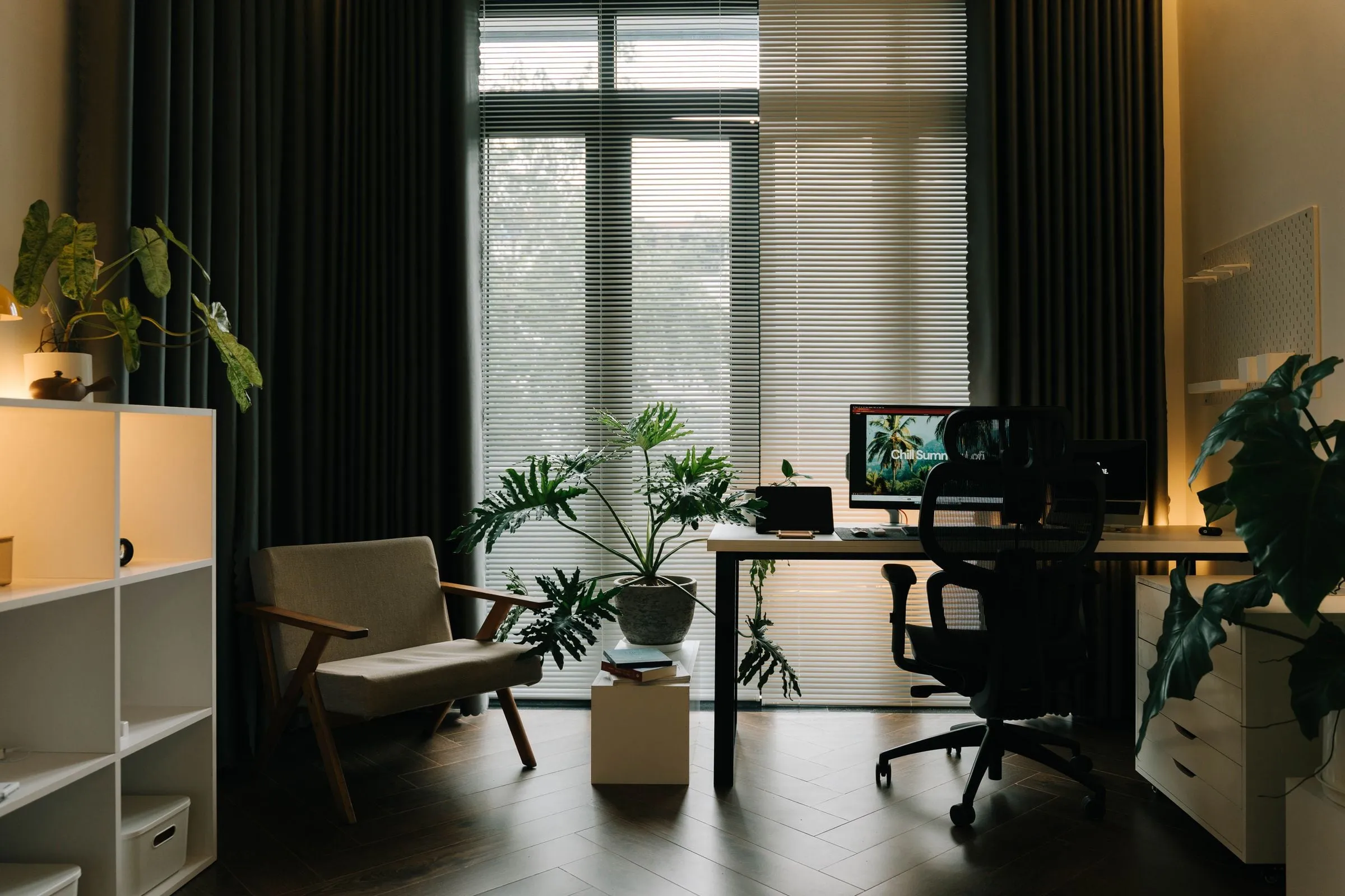 A modern office with a standing desk, ergonomic chair, lounge chair and plants by tall windows